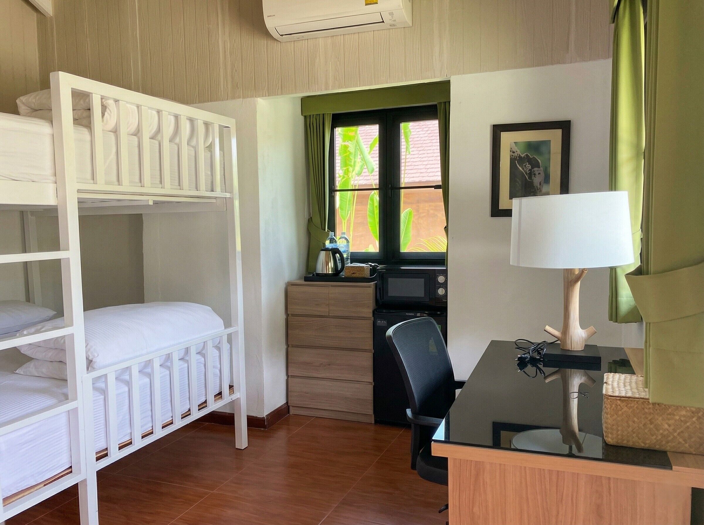 A room with bunk beds, a desk with a chair, a microwave, a small cabinet, and a window with curtains.