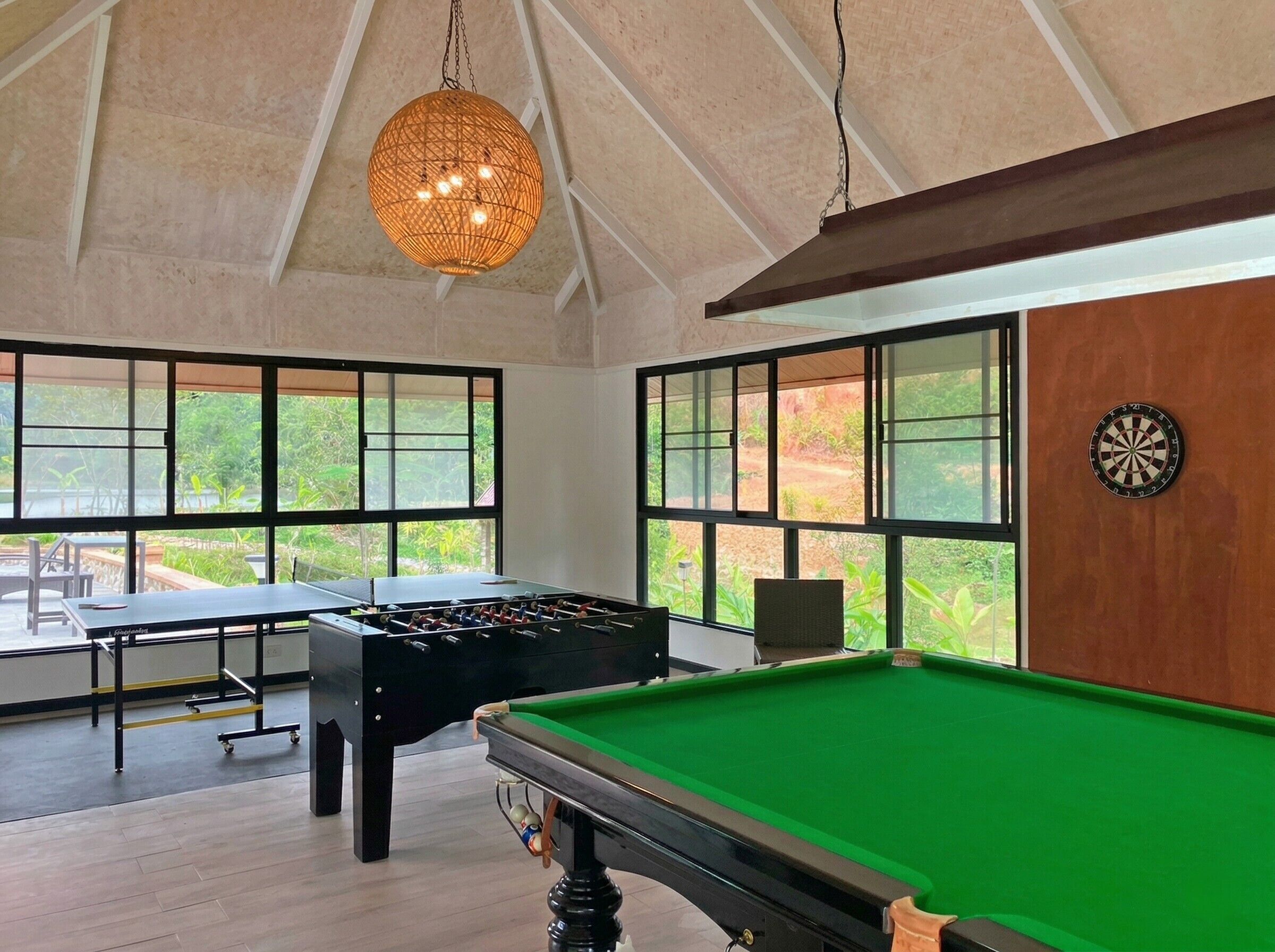 A pool table, foosball table, and dartboard in a room with large windows and a hanging light fixture.