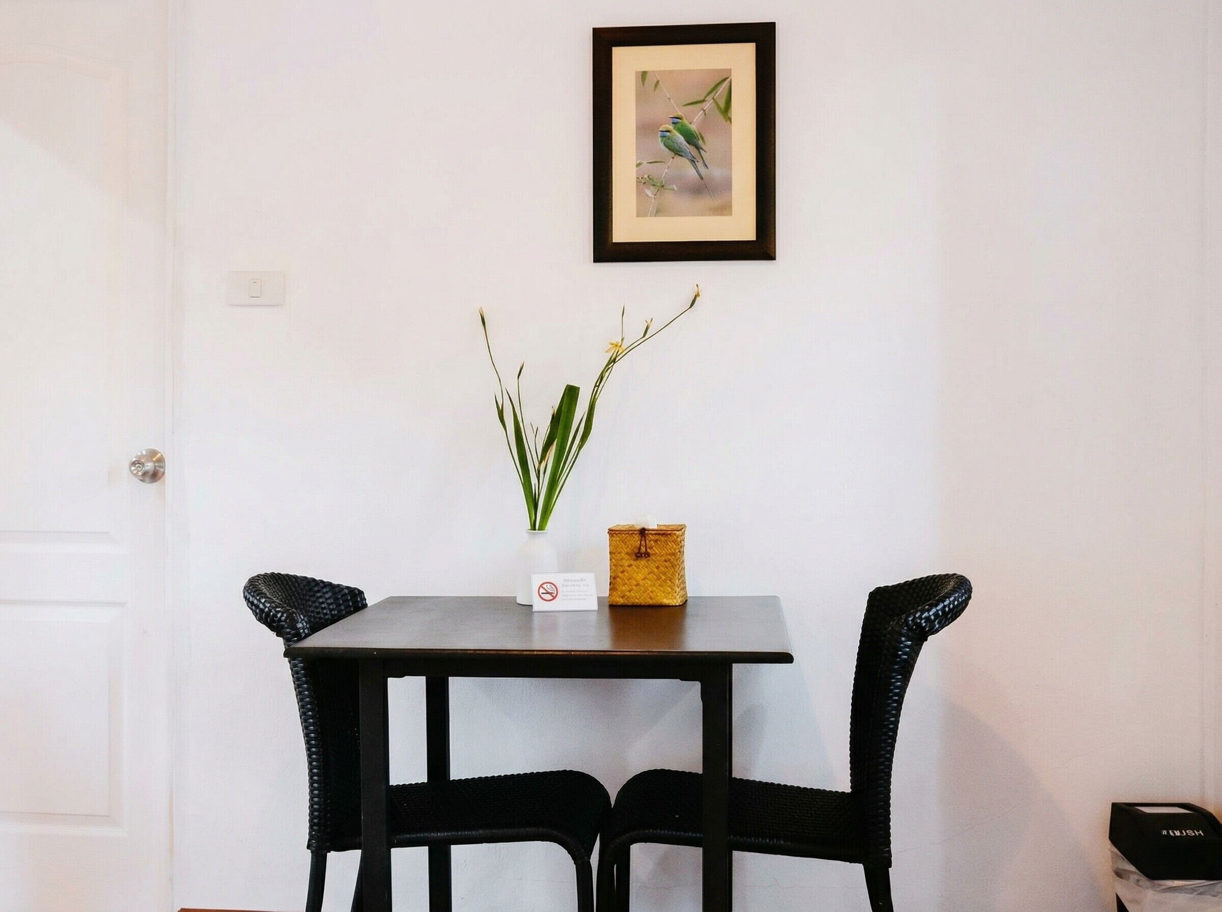 A small dining area with a table and two chairs, a framed picture on the wall, and a vase with plants.