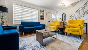 Een woonkamer met een blauwe bank, gele fauteuils, een salontafel en een trap met een decoratieve balustrade.