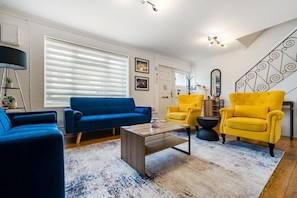 A living room with a blue sofa, yellow armchairs, a coffee table, and a staircase with a decorative railing. - Free parking, private garden and Elizabeth Line at your door (Greater London)