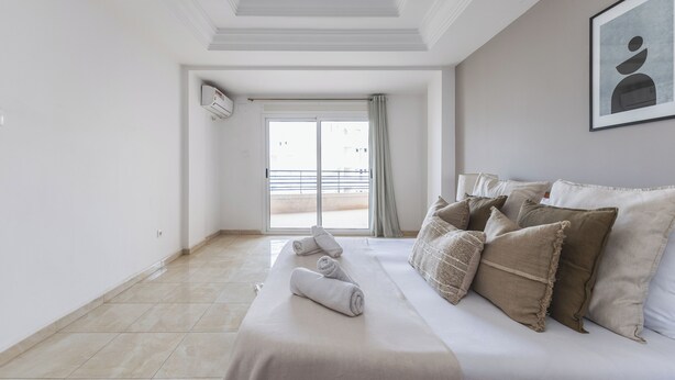 A hotel room with a bed, pillows, a towel, a framed picture, and a view through a door.