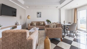 A modern living room with a sofa set, a coffee table, a TV mounted on the wall, and a dining area with a table and chairs.