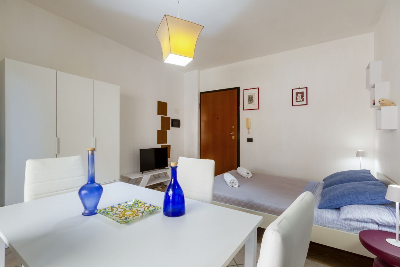 Una habitación de hotel moderna con una mesa blanca, dos jarrones azules, un televisor, una cama con almohadas azules, una puerta de madera y un pequeño estante con un osito de peluche.