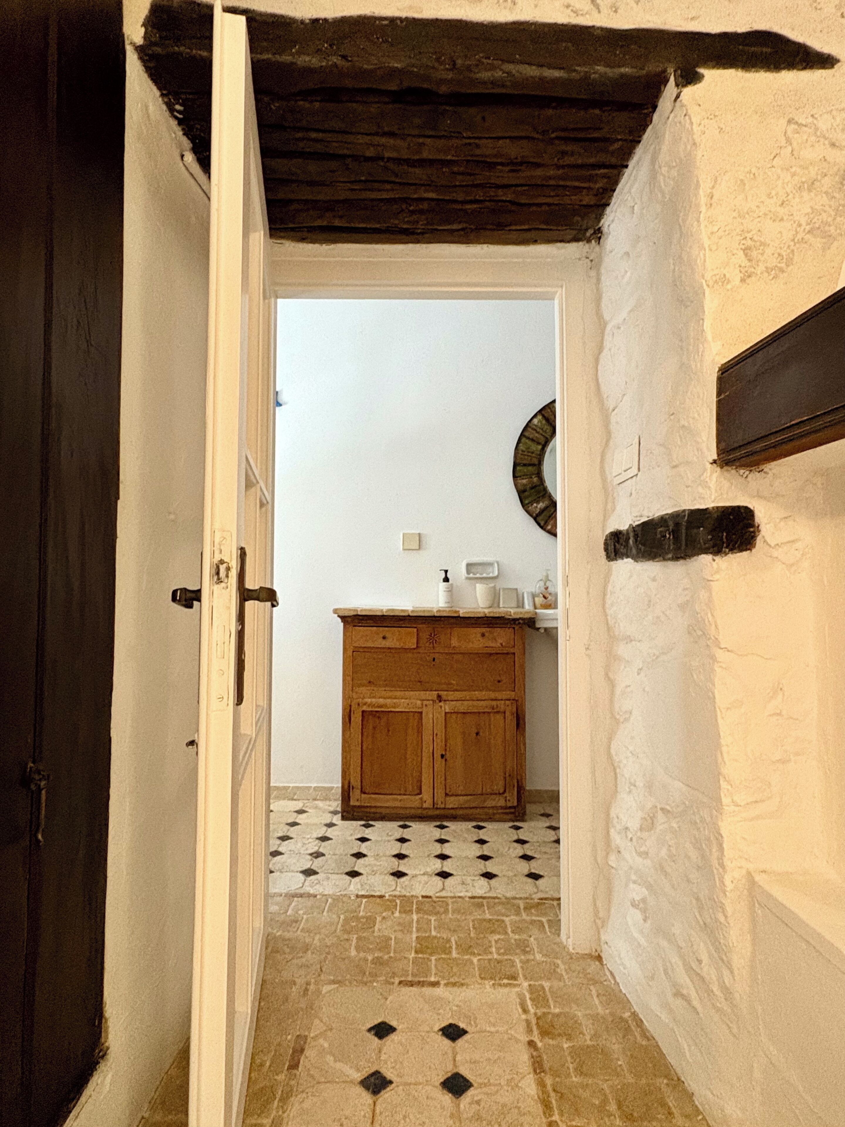 A narrow hallway with a wooden door on the left, a tiled floor, a wooden cabinet with a mirror above it, and a white wall.