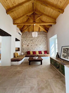 A spacious living area with a stone wall, wooden ceiling beams, a sofa with cushions, a coffee table, and a decorative cabinet. - Siena House Hydra - Renovated in 2026 (Idra)