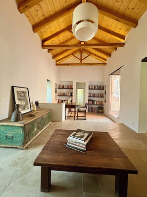 A spacious room with a high wooden ceiling, a central table with books, a bookshelf, and a door leading to another room. - Siena House Hydra - Renovated in 2026 (Idra)