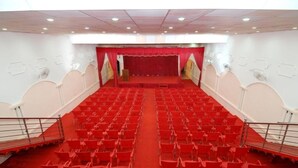 Un amplio auditorio con filas de sillas rojas, un escenario con cortinas rojas y una pared blanca con detalles decorativos.