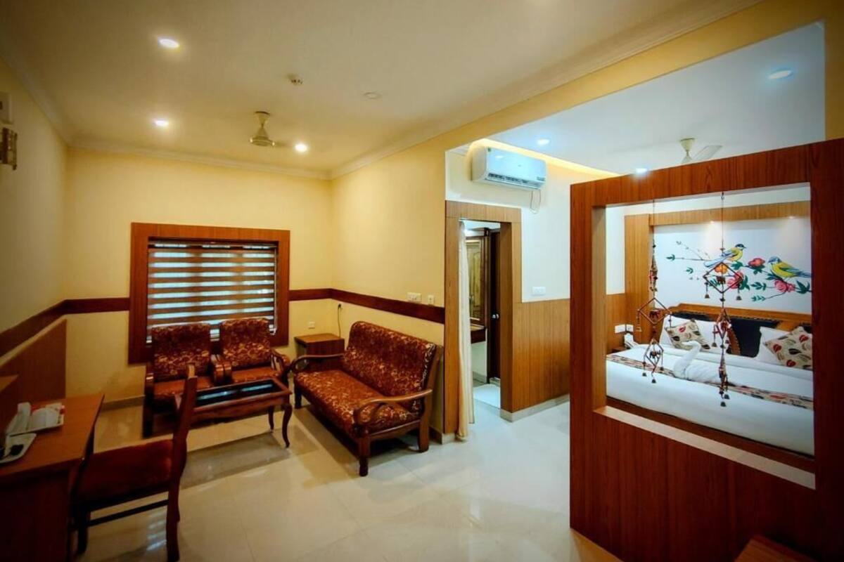 A hotel room with a sofa set, a dining table, a small kitchen area, and a decorative window with a floral design.