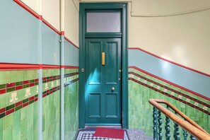 Eine grüne Tür mit einem rot-weiß gestreiften Sockel, eine rote Fußmatte am Fuß einer Treppe und eine Wand mit grünen und roten Fliesen.