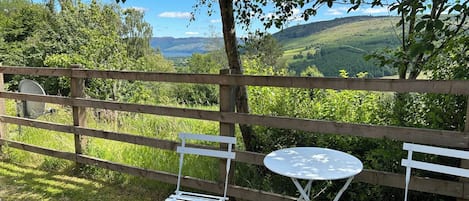 Une clôture en bois, deux chaises en métal blanches et une petite table ronde, le tout installé dans un espace verdoyant offrant une vue sur des collines et des arbres.