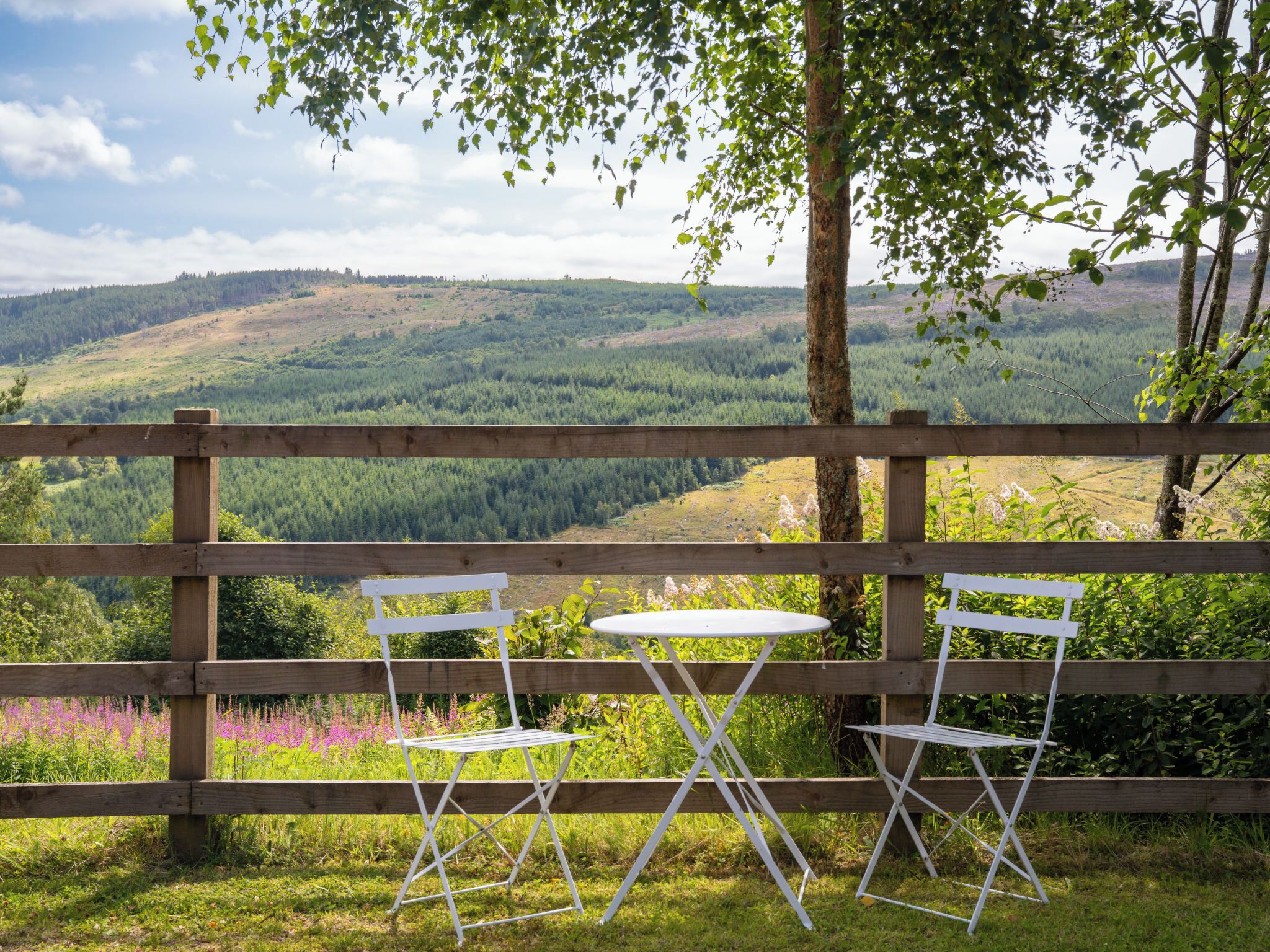A wooden fence with a table and two chairs in a grassy area, overlooking a forested valley.