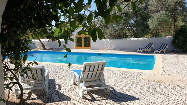 En swimmingpool med solstolar, en stenbelagd uteplats och en vit vägg med en dörr.