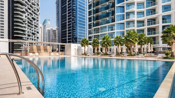 A swimming pool with a tiled deck, surrounded by lounge chairs and umbrellas, with modern buildings in the background. - Gala Guests - Maison Prive Collection (Dubai)