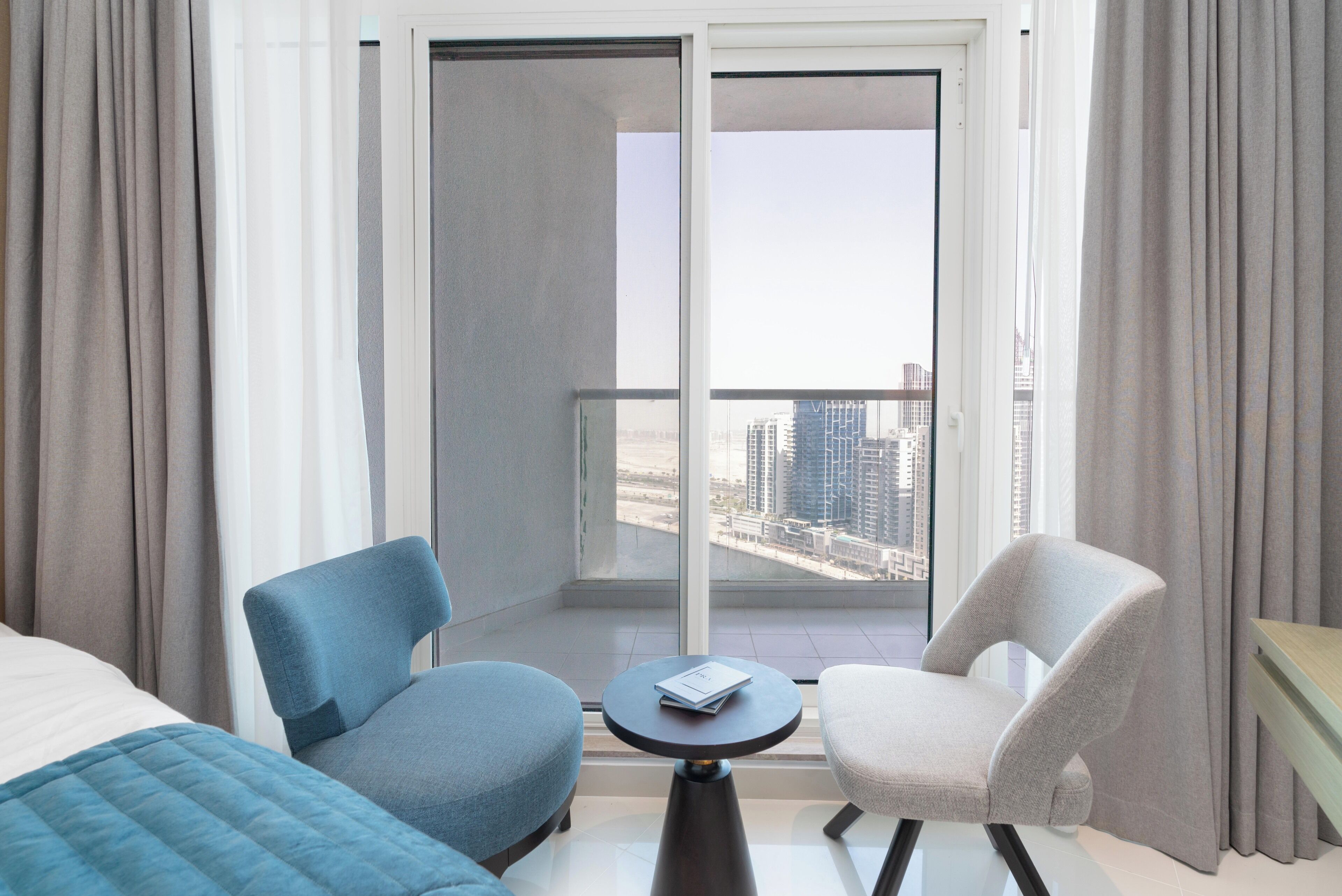Une chambre d’hôtel avec un lit, deux chaises, une petite table et une vue sur le paysage urbain, visible à travers une grande fenêtre.