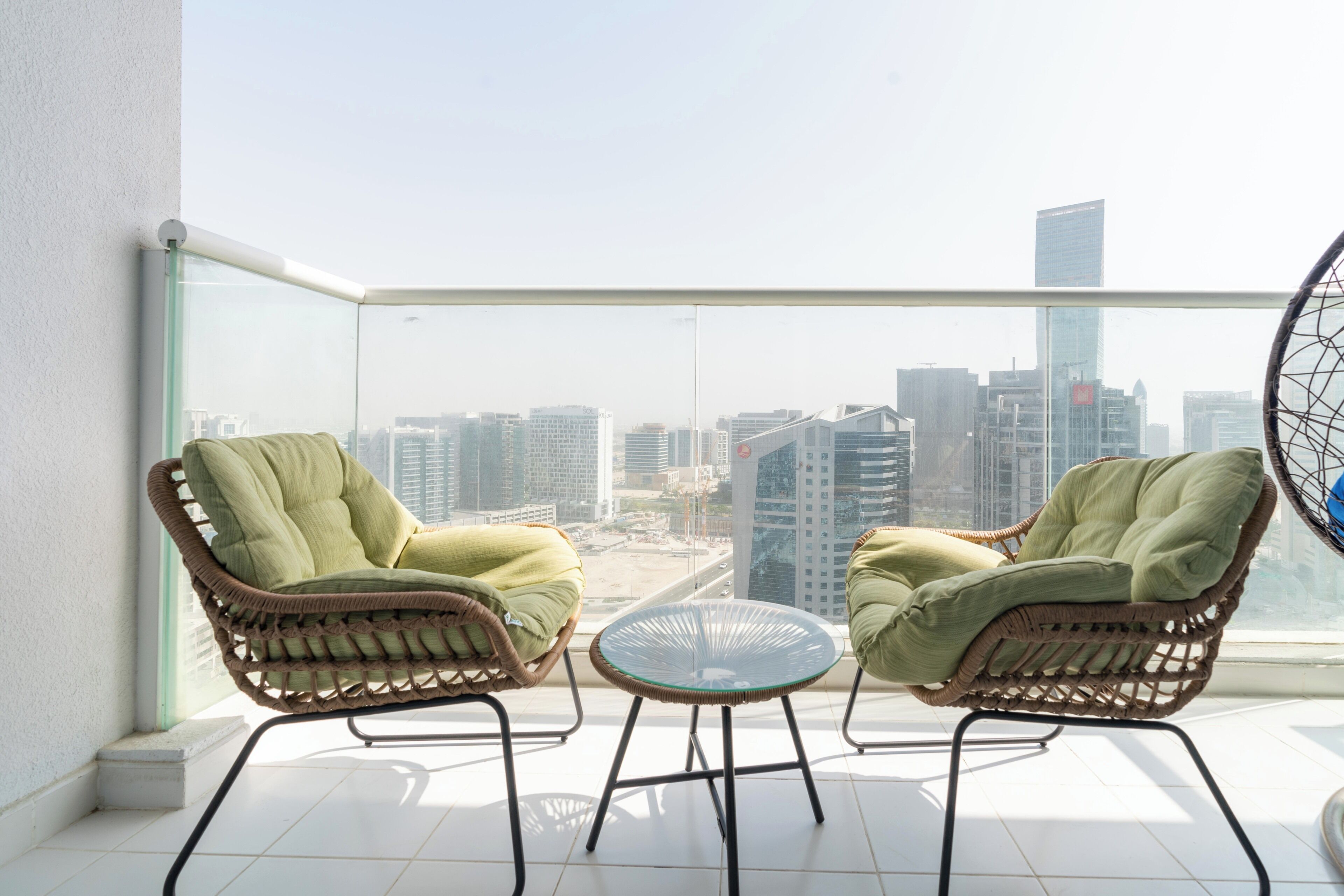 A balcony with two wicker chairs and a small table, overlooking a cityscape with modern buildings.
