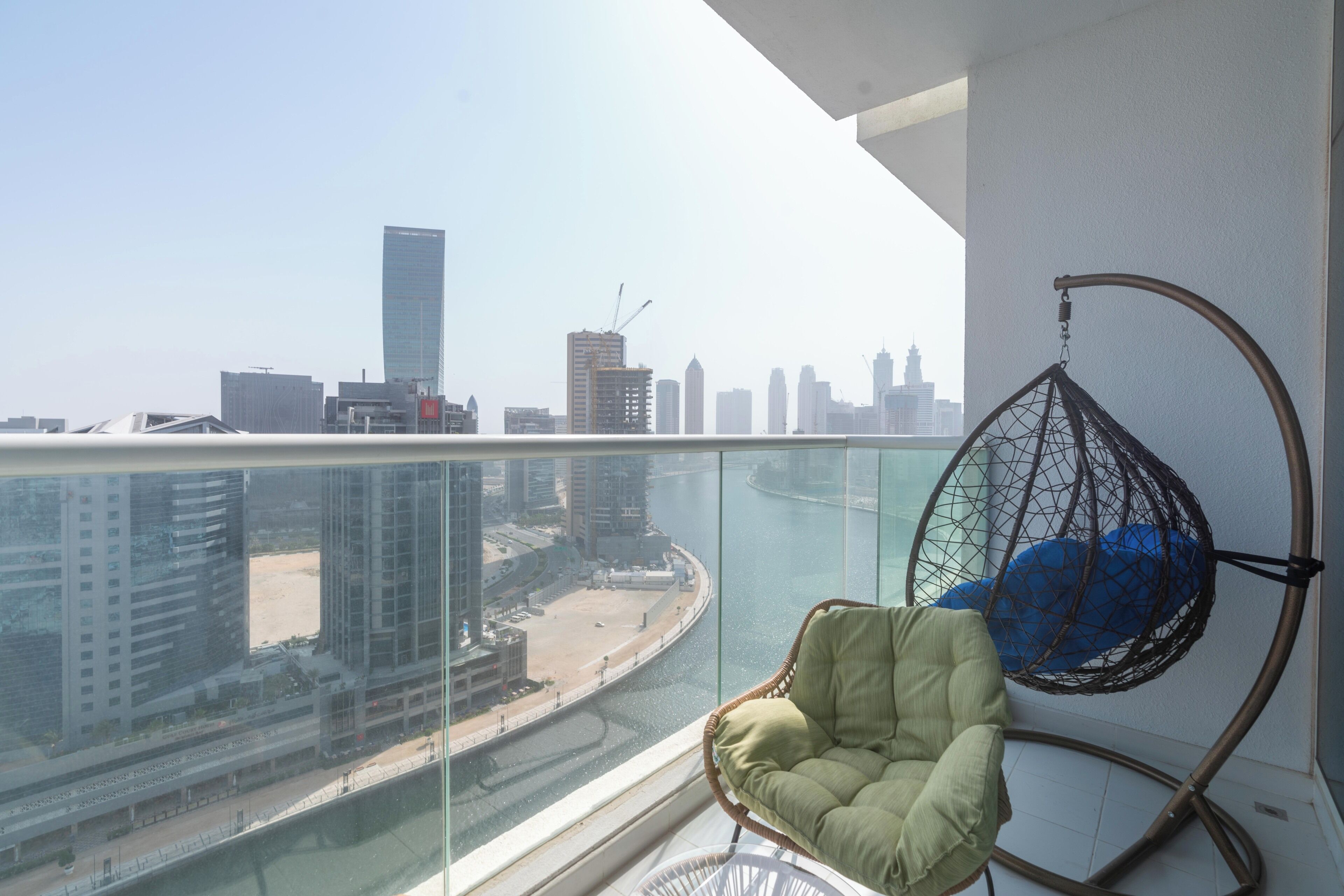A balcony with a hanging chair, a cushioned chair, and a view of a cityscape with buildings and a waterfront.