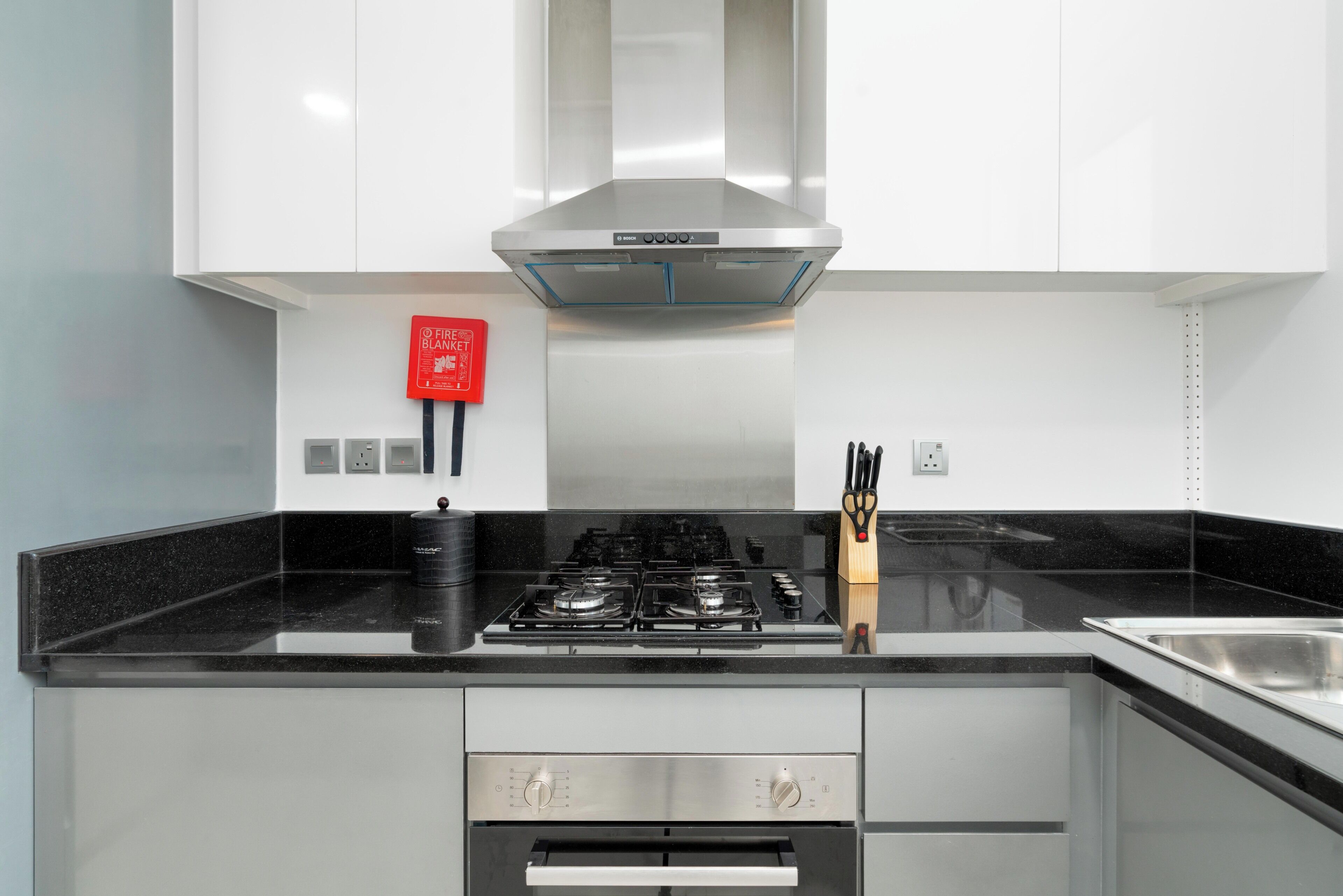 A modern kitchen with a stainless steel range hood, a gas stove, a built-in oven, and a red fire extinguisher on the wall.