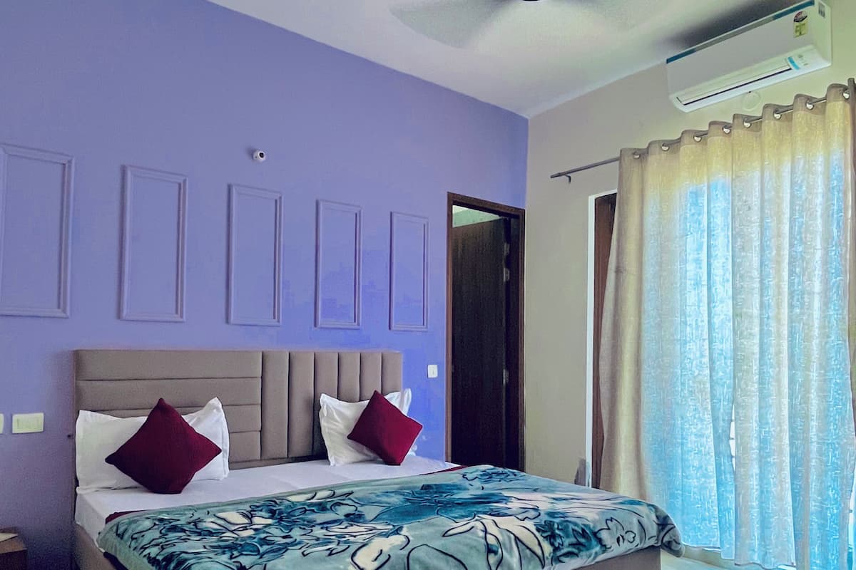 Un dormitorio con cama, ventilador de techo, equipo de aire acondicionado, ventana con cortinas y una puerta.