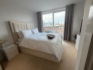 A modern bedroom with a large bed, a nightstand, a dresser, and a view of a cityscape through a large window. - Apartment Sleeps 4 With Dishwasher (London)