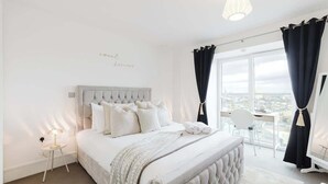 A bedroom with a large bed, a bedside table with a lamp, a view of the city through the window, and a balcony with a chair. - Luxury 1BR w/City views & A/C - Clapham (Greater London)