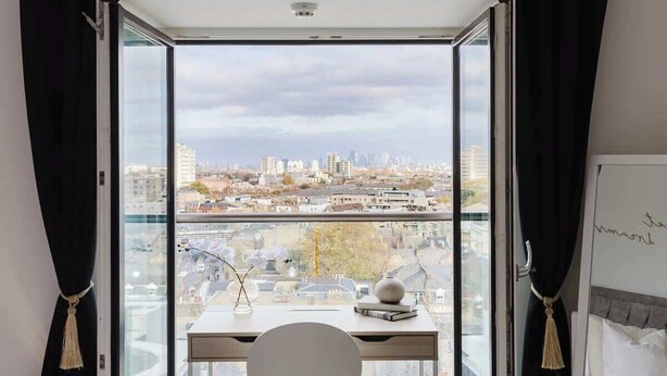 A room with a desk, a chair, a view of a cityscape through a large window, and a whiteboard with writing. - Luxury 1BR w/City views & A/C - Clapham (Greater London)