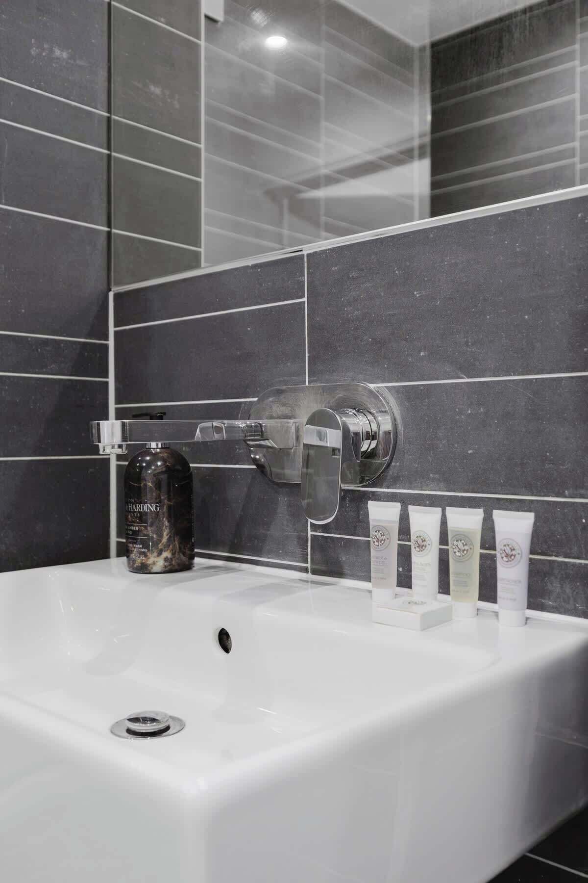 Un baño moderno con un lavamanos blanco, paredes de azulejos oscuros, un grifo cromado y una botella de jabón para manos.