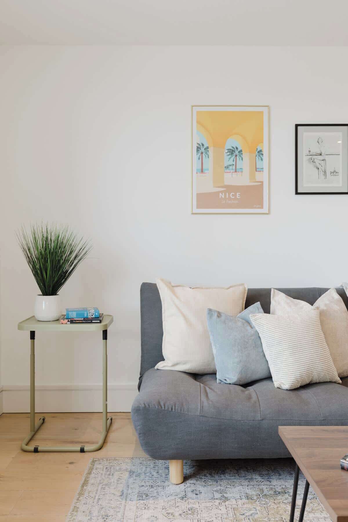 Un salon avec un canapé gris, une petite table d’appoint sur laquelle est posée une plante, et un tableau encadré accroché au mur.