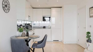A modern kitchen with white cabinets, a round dining table with two chairs, and a clock on the wall. - Luxury 1BR w/City views & A/C - Clapham (Greater London)