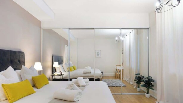 Une chambre d’hôtel moderne avec un grand lit, une cloison en verre, une petite table avec une lampe et une plante en pot.