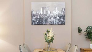 Un coin repas comprenant une table, des chaises, un vase avec des fleurs et un tableau représentant un paysage urbain accroché au mur.