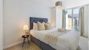 Quarto com cama grande, abajur e vista para edifícios pela janela.