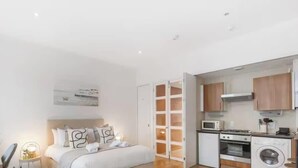 Ett sovrum med en säng, ett sängbord och ett litet bord. Det finns ett litet kök med mikrovågsugn, ugn och tvättmaskin.