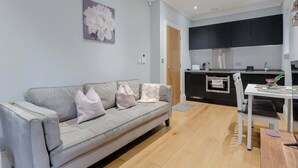 A modern living room with a grey sofa, a dining table with chairs, a kitchen area with black cabinets and stainless steel appliances, and a door leading to another room. - Delightful 1Bed-Private Patio-FR Parking-PingPong (Greater London)