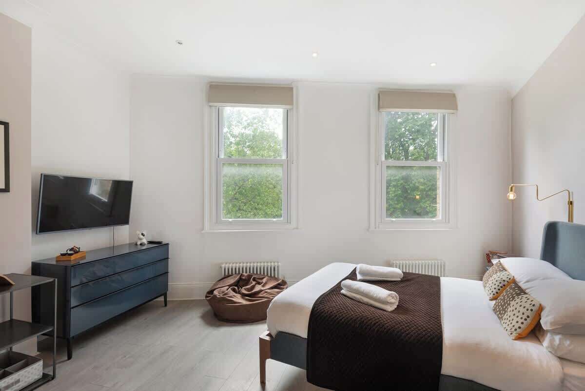 A modern bedroom with a bed, a TV, a dresser, and two windows with a view of greenery.