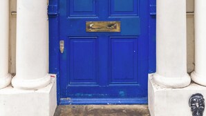 Een blauwe deur met een messing klink, omringd door witte zuilen, en een klein raam erboven.