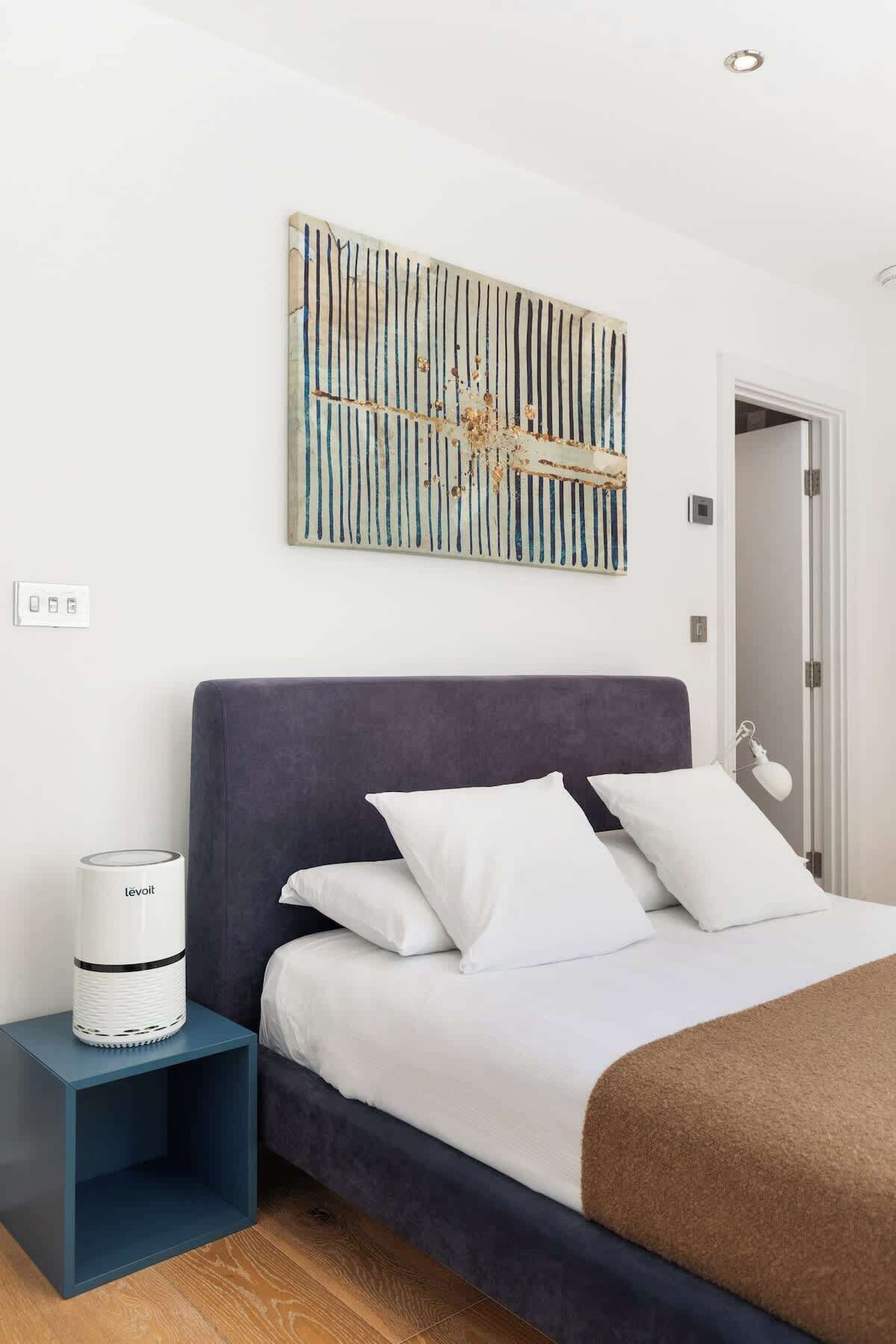 A bedroom with a bed, white pillows, a dark headboard, a bedside table with a white lamp, and a wall-mounted abstract painting.