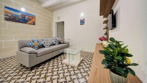 Sala de estar com sofá, almofadas estampadas, mesa de centro de vidro, planta em vaso e TV fixada na parede.