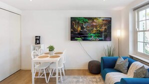 A modern living room with a dining area, a blue sofa, and a TV displaying a nature scene. - Luxe 1BD + A/C | King Bed & 75" TV | Notting Hill (Greater London)