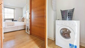 A laundry room with a washing machine, a wooden door, and a white wall. - Luxe 1BD + A/C | King Bed & 75" TV | Notting Hill (Greater London)
