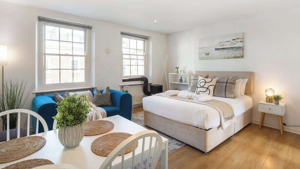 A modern bedroom with a bed, bedside tables, a window, and a dining area with a table and chairs. - Luxe 1BD + A/C | King Bed & 75" TV | Notting Hill (Greater London)