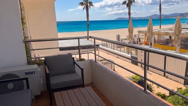 Ein Balkon mit Tisch und Stühlen mit Blick auf einen Strand, gesäumt von Palmen und Sonnenschirmen.