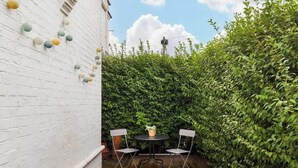 Eine Terrasse mit einem runden Tisch und zwei Stühlen, umgeben von einer weißen Ziegelmauer und einer Hecke.