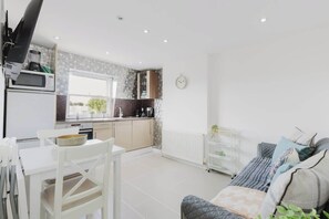 Eine moderne Küche mit weißen Geräten, ein Essbereich mit weißem Tisch und Stühlen sowie eine Wohnbereich mit einer Couch und Kissen.