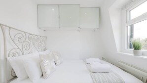 Una camera da letto con un letto, biancheria da letto bianca, una testiera decorativa, una finestra con una pianta e un mobiletto a parete.