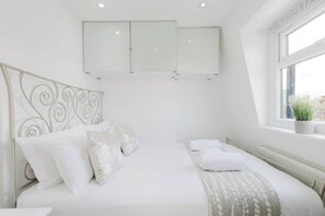 Ein Schlafzimmer mit Bett, weißer Bettwäsche, einem dekorativen Kopfteil, einem Fenster mit Pflanze und einem an der Wand befestigten Schrank.
