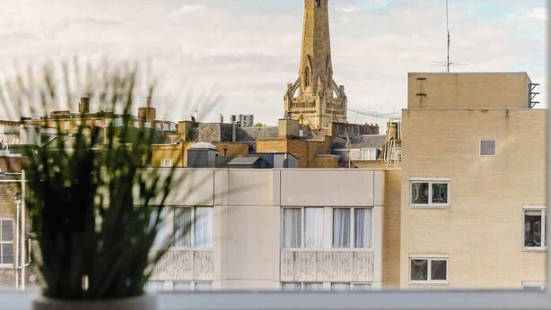 Vista dalla finestra su una città, con una guglia di chiesa ben visibile, una pianta in vaso sul davanzale e edifici moderni in primo piano.