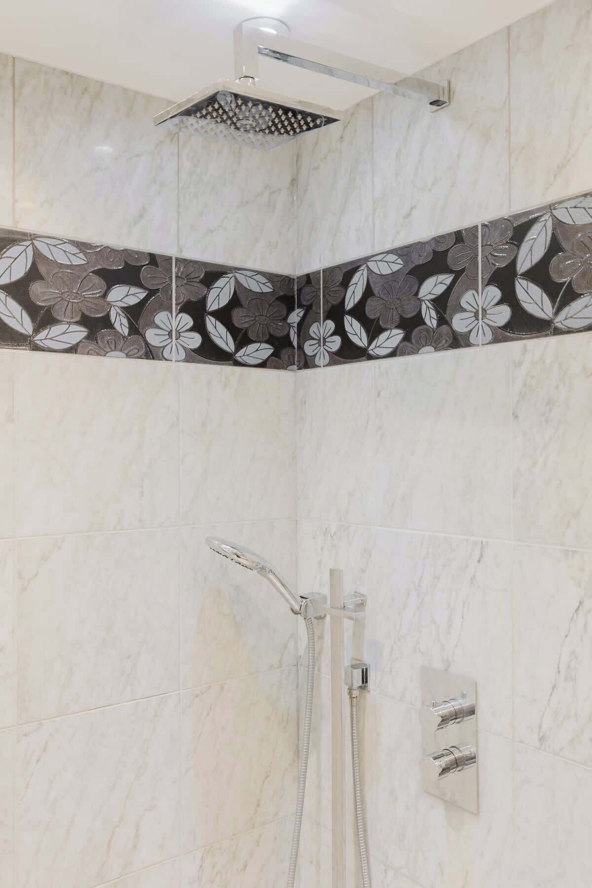 Ducha con cabezal de ducha lluvia y cabezal de ducha de mano, diseño de azulejos en esquina con motivos florales y pared de mármol.
