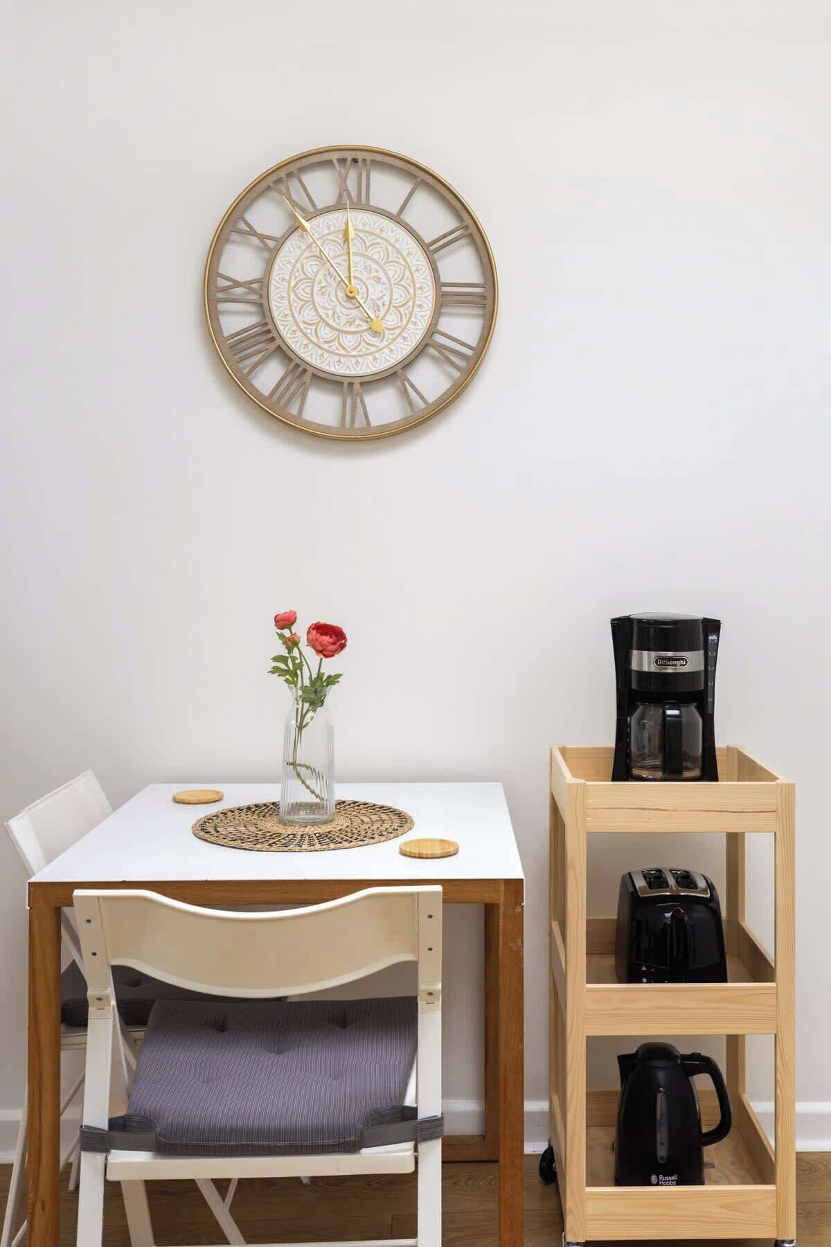 Una mesa de comedor con un reloj, un jarrón con flores y una cafetera en un estante.