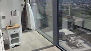Un dormitorio moderno con un ventanal grande, una mesita de noche con lámpara y vistas a un paisaje urbano.